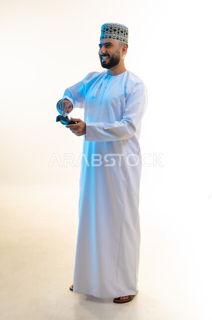 قضاء وقت ممتع ومسلي، اجهزة وتقنيات الكترونية ترفيهيه حديثة, التفاعل والاندماج مع ألعاب الفيديو، بورتريه لشاب عربي خليجي عماني يرتدي الدشداشة والكمة يقوم بالضغط على ازرار عصا التحكم، صورة لكامل الجسم، خلفية بيضاء