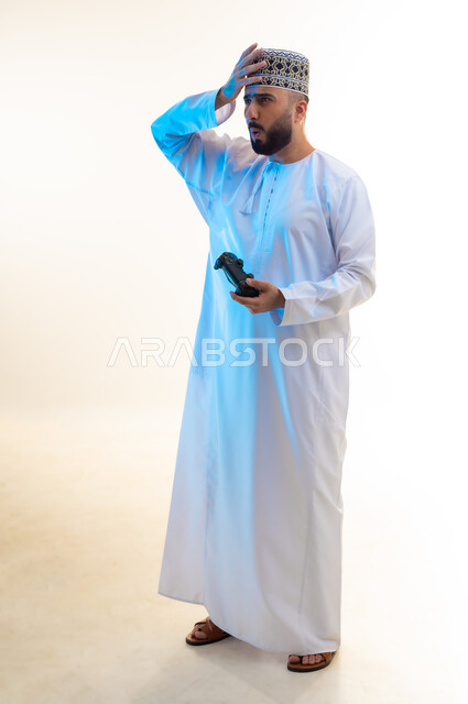 Interacting and merging with video games, having a fun and entertaining time, modern entertainment electronic devices and technologies, portrait of a young Gulf Arab Omani man wearing a dishdasha and a turban, holding a joystick, putting his hand on his head with gestures of disappointment, full body image, white background