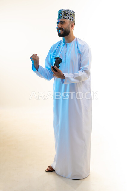 Raising fist with victory gestures, having fun playing video games, enjoying modern technology and techniques, concept of fun and entertainment, portrait of an Arab Gulf Omani man wearing a dishdasha and a kummah holding a joystick in his hand, full body, white background