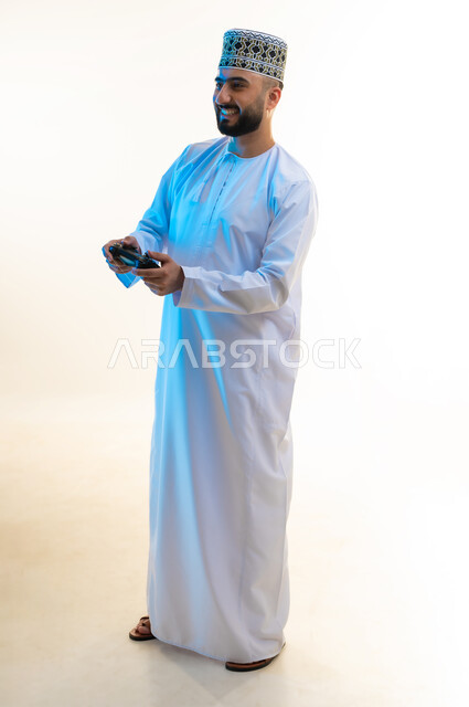 قضاء أوقات ممتعة في اللعب بألعاب الفيديو، الاستمتاع بالتقنيات والتكنولوجيا الحديثة، مفهوم المتعة والتسلية، إيماءات الاندماج والتفاعل, بورتريه لرجل عربي خليجي عماني يرتدي الدشداشة والكمة يمسك بيده عصا التحكم، الجسم بالطول الكامل، خلفية بيضاء