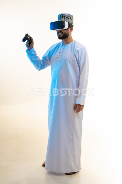 محاكاة العالم الخيالي، استخدام الأجهزة التكنولوجية والتقنيات الحديثة، بورتريه لرجل عربي خليجي عماني يرتدي الدشداشة والكمة ونظارة الواقع الإفتراضي يمسك بيده عصا التحكم يقوم بلعب ألعاب الفيديو الترفيهية بإيماءات الاستمتاع والحماس، الجسم بالطول الكامل، خلفية بيضاء