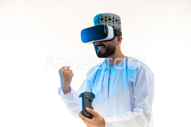 استخدام أجهزة تكنولوجية وتقنيات حديثة، بورتريه مقرب لرجل عربي خليجي عماني يرتدي الدشداشة والكمة ونظارة الواقع الإفتراضي يمسك عصا التحكم، رفع قبضة اليد بايماءات الانتصار، دمج العالم الخيالي مع المتعة والتسلية، اللعب بألعاب الفيديو الترفيهية، خلفية بيضاء