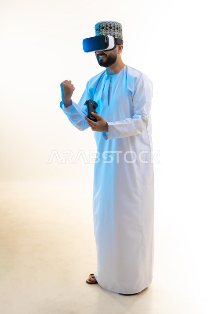 أجهزة تكنولوجية وتقنيات حديثة، بورتريه لرجل عربي خليجي عماني يرتدي الدشداشة والكمة ونظارة الواقع الإفتراضي يمسك عصا التحكم، رفع قبضة اليد بايماءات الانتصار، دمج العالم الخيالي مع المتعة والتسلية، اللعب بألعاب الفيديو الترفيهية، الجسم بالطول الكامل، خلفية بيضاء