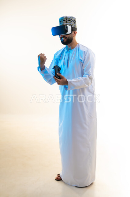 دمج العالم الخيالي مع المتعة والتسلية، أجهزة تكنولوجية وتقنيات حديثة، بورتريه لرجل عربي خليجي عماني يرتدي الدشداشة والكمة ونظارة الواقع الإفتراضي يمسك عصا التحكم، رفع قبضة اليد بايماءات الانتصار، اللعب بألعاب الفيديو الترفيهية، الجسم بالطول الكامل، خلفية بيضاء