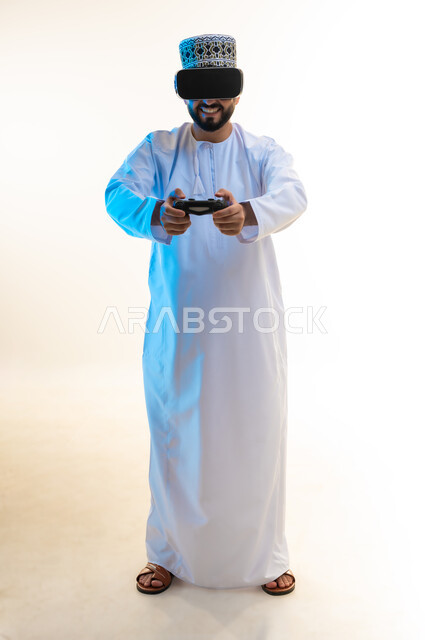 دمج العالم الخيالي مع المتعة والتسلية، اللعب بألعاب الفيديو الترفيهية، استخدام الأجهزة التكنولوجية والتقنيات الحديثة، بورتريه لرجل عربي خليجي عماني يرتدي الدشداشة والكمة ونظارة الواقع الإفتراضي يمسك بيده عصا التحكم، الجسم بالطول الكامل، خلفية بيضاء
