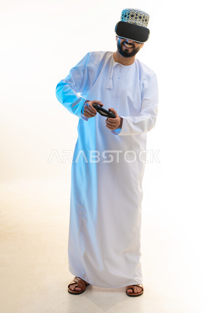 اللعب بألعاب الفيديو الترفيهية، استخدام الأجهزة التكنولوجية والتقنيات الحديثة، بورتريه لرجل عربي خليجي عماني يرتدي الدشداشة والكمة ونظارة الواقع الإفتراضي يمسك بيده عصا التحكم، دمج العالم الخيالي مع المتعة والتسلية، الجسم بالطول الكامل، خلفية بيضاء