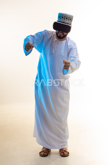 تقنيات وأجهزة تكنولوجية، محاكاة ركوب دراجة ثري دي في العالم الخيالي، بورتريه لرجل عربي خليجي عماني يرتدي الدشداشة والكمة ونظارة الواقع الإفتراضي ثلاثية الأبعاد يقوم باللعب في ألعاب الفيديو، إيماءات السعادة بألعاب قيادة الدراجات، الجسم بالطول الكامل، خلفية بيضاء