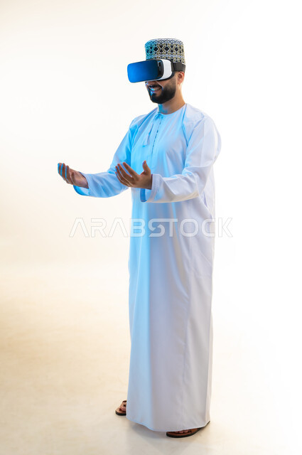 التطور التقني والتكنولوجي، إمساك وحمل عنصر ما، بورتريه لرجل عربي خليجي عماني يرتدي الدشداشة والكمة يضع نظارة ثلاثية الأبعاد يرفع يديه للأعلى، الاندماج ومحاكاة العالم الخيالي, مشاهدة الأفلام عبر تقنيات الواقع الافتراضي, الجسم بالطول الكامل، خلفية بيضاء