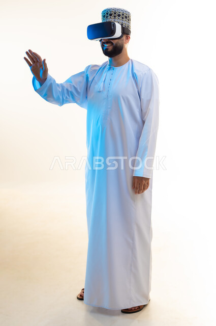 مشاهدة الأفلام عبر تقنيات الواقع الافتراضي, رفع اليدين بإيماءات الاندماج والاستمتاع، محاولة لمس عنصر ما، محاكاة العالم الخيالي, بورتريه من الجانب لرجل عربي خليجي عماني يرتدي الدشداشة والكمة يضع نظارة ثلاثية الأبعاد يتفاعل معها، الجسم بالطول الكامل، خلفية بيضاء