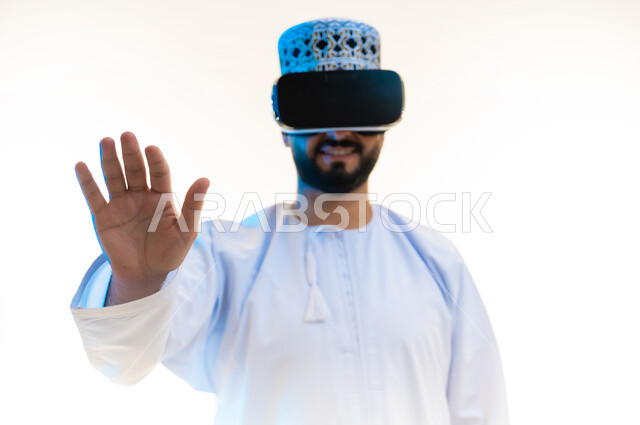 Raising hands with gestures of integration and enjoyment, trying to touch an item, simulating a fantasy world, close-up portrait of an Arab Gulf Omani man wearing a dishdasha and a kuma wearing 3D glasses and interacting with them, watching movies via virtual reality technologies, white background