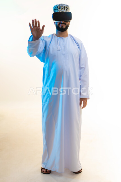 محاولة لمس عنصر ما، محاكاة العالم الخيالي, بورتريه لرجل عربي خليجي عماني يرتدي الدشداشة والكمة يضع نظارة ثلاثية الأبعاد يقوم بالتفاعل معها، رفع اليدين بإيماءات الاندماج والاستمتاع، مشاهدة الأفلام عبر تقنيات الواقع الافتراضي, الجسم بالطول الكامل، خلفية بيضاء