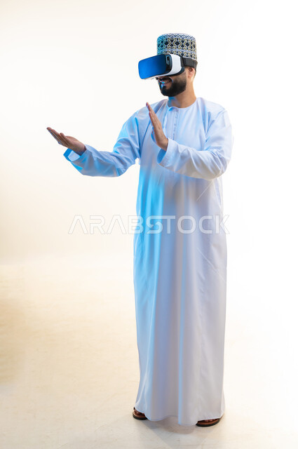 رفع اليدين بإيماءات الاندماج والاستمتاع، مشاهدة الأفلام باستخدام تقنيات الواقع الافتراضي, محاكاة العالم الخيالي, بورتريه لرجل عربي خليجي عماني يرتدي الدشداشة والكمة يضع نظارة ثلاثية الأبعاد يقوم بالتفاعل معها، الجسم بالطول الكامل، خلفية بيضاء