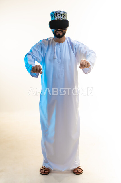 تقنيات وأجهزة تكنولوجية، إيماءات السعادة بألعاب قيادة الدراجات، محاكاة ركوب دراجة ثري دي في العالم الخيالي، بورتريه لرجل عربي خليجي عماني يرتدي الدشداشة والكمة ونظارة الواقع الإفتراضي ثلاثية الأبعاد يقوم باللعب في ألعاب الفيديو، الجسم بالطول الكامل، خلفية بيضاء