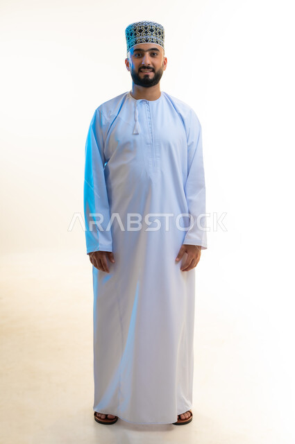 الاهتمام بالمظهر الخارجي والأناقة، مفهوم الرجولة والوسامة، الوقوف بشكل مستقيم بإيماءات الثقة بالنفس، بورتريه لشاب عربي خليجي عماني يرتدي الدشداشة والكمة ينظر إلى الكاميرا بتعابير السرور، الجسم بالطول الكامل، خلفية بيضاء