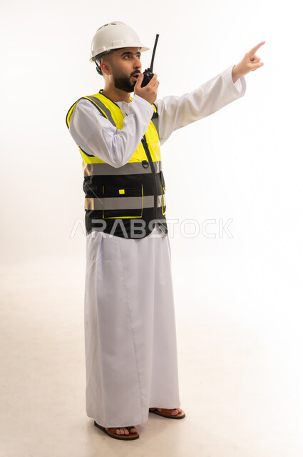 المتابعة والإشراف على المشاريع الهندسية، بورتريه لمهندس عربي خليجي عماني يرتدي الدشداشة والخوذة وسترة الحماية يحمل جهاز اتصال لاسلكي يشير بإصبع السبابة إلى شيء ما في الأعلى، مهن ووظائف شبابية، إعطاء أوامر خاصة بالعمل، صورة الجسم بالطول الكامل، خلفية بيضاء