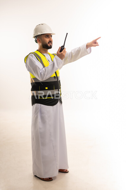 إدارة المشاريع الهندسية الإعمارية، بورتريه لمهندس عربي خليجي عماني يرتدي الدشداشة والخوذة وسترة الحماية يحمل جهاز اتصال لاسلكي يشير بإصبع السبابة إلى شيء ما في الأعلى، المهن والوظائف الشبابية، إعطاء أوامر خاصة بالعمل، صورة الجسم بالطول الكامل، خلفية بيضاء