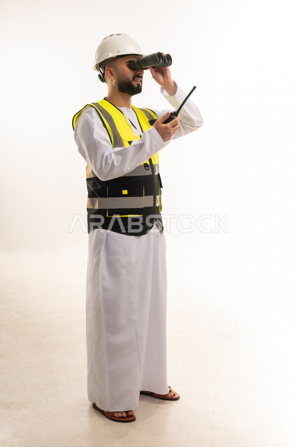 Youth jobs and professions, following up on engineering projects, seeing distant objects, portrait of an Arab Gulf Omani engineer wearing a dishdasha, helmet and protective vest, holding a wireless communication device, looking through binoculars, giving work orders, full-length body image, white background