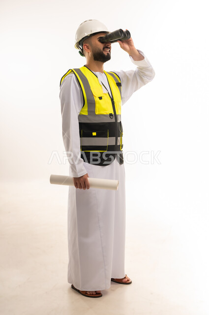 رؤية الأشياء البعيدة، بورتريه لمهندس عربي خليجي عماني يرتدي الدشداشة والخوذة وسترة الحماية ينظر من خلال المنظار ثنائي العينين إلى شيء ما يحمل بيده مخطط، متابعة سير المشاريع الهندسية الإعمارية، المهن والوظائف الشبابية، صورة الجسم بالطول الكامل، خلفية بيضاء