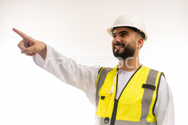 المهن والوظائف الشبابية، مفهوم الإنشاءات المعمارية والهندسة، بورتريه مقرب لمهندس عربي خليجي عماني مبتسم يرتدي الدشداشة والخوذة وسترة الحماية يشير بإصبع السبابة إلى شيء ما في الأعلى، متابعة سير المشاريع الإعمارية، خلفية بيضاء