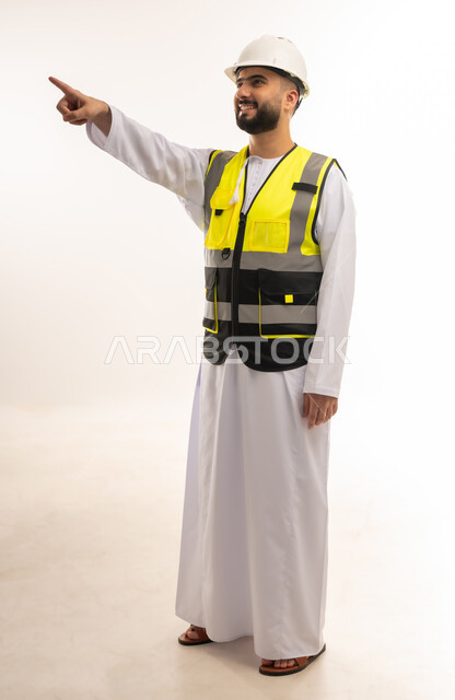 متابعة سير المشاريع الإعمارية، المهن والوظائف الشبابية، مفهوم الإنشاءات المعمارية والهندسة، بورتريه لمهندس عربي خليجي عماني يرتدي الدشداشة والخوذة وسترة الحماية يشير بإصبع السبابة إلى شيء ما في الأعلى، صورة الجسم بالطول الكامل، خلفية بيضاء