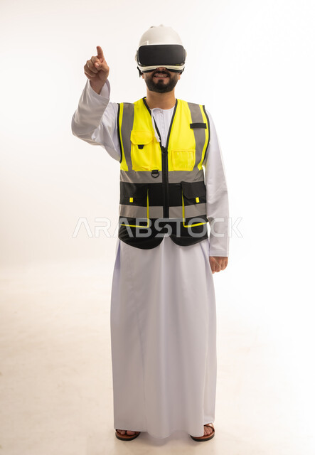 رفع اصبع السبابة في محاولة للمس العناصر الهولوجرامية، استخدام التقنيات الحديثة والمتطورة في مجال التصاميم المعمارية، بورتريه لمهندس عربي خليجي عماني يرتدي الدشداشة والسترة وخوذة الحماية يلبس نظارات الفي آر ثلاثية الأبعاد، الجسم بالطول الكامل، خلفية بيضاء