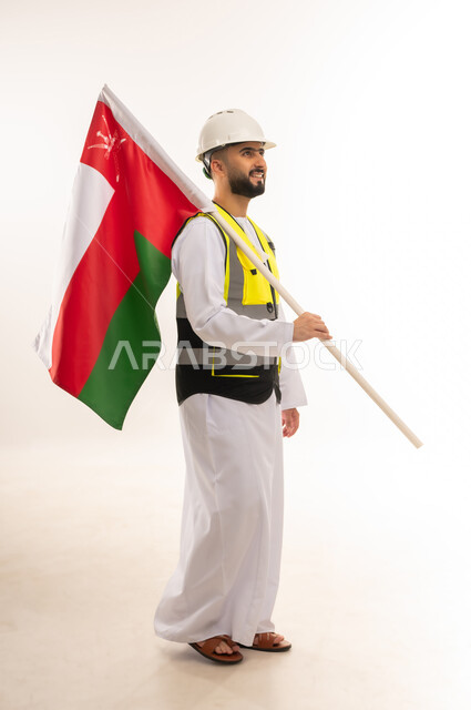 الاعتزاز بالراية العمانية، المشاركة بالاحتفال بذكرى اليوم الوطني، إيماءات الفخر بالانتماء للبلاد، بورتريه من الجانب لمهندس عربي خليجي عماني يرتدي الدشداشه والسترة وخوذة الحماية يحمل علم السلطنة على كتفه، الجسم بالطول الكامل، خلفية بيضاء