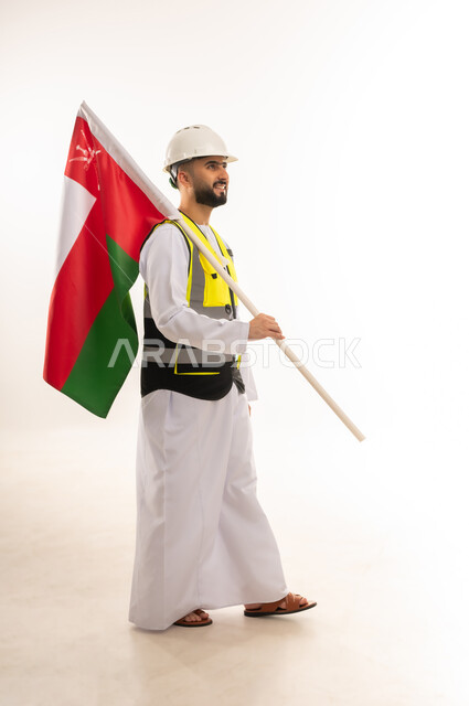 Participating in the celebration of the National Day, gestures of pride in belonging to the country, pride in the Omani flag, side portrait of an Arab Gulf Omani engineer wearing a dishdasha, jacket and protective helmet, carrying the Sultanate's flag on his shoulder, full-length body, white background