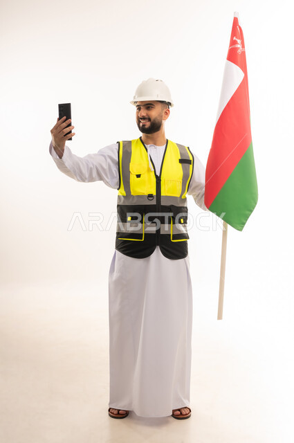 التقاط صور السيلفي التذكارية باستخدام الجوال، الاعتزاز بالراية العمانية، المشاركة بالاحتفال بذكرى اليوم الوطني، الفخر بالانتماء للبلاد، بورتريه لمهندس عربي خليجي عماني يرتدي الدشداشه والسترة وخوذة الحماية يحمل علم سلطنة عمان، الجسم بالطول الكامل، خلفية بيضاء