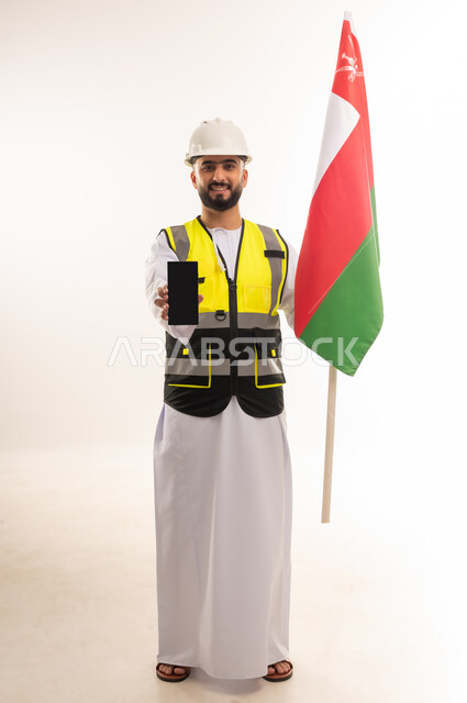 عرض شاشة جوال سوداء فارغة، الاعتزاز بالراية العمانية، المشاركة بالاحتفال بذكرى اليوم الوطني، إيماءات الفخر بالانتماء للبلاد، بورتريه لمهندس عربي خليجي عماني يرتدي الدشداشه والسترة وخوذة الحماية يحمل علم سلطنة عمان بثقة بالنفس، الجسم بالطول الكامل، خلفية بيضاء