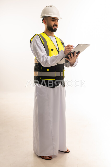 إدارة الأعمال والاطلاع على آخر المستجدات، تدوين وتسجيل الملاحظات الهامة، بورتريه لمهندس عربي خليجي عماني يرتدي الدشداشة والسترة وخوذة الحماية يمسك ملف بيده ويكتب فيه، التأكد من سير العمل بالشكل المطلوب والصحيح، الجسم بالطول الكامل، خلفية بيضاء