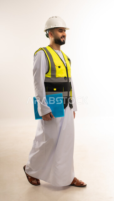 إيماءات السير والتقدم نحو الأمام، إدارة الأعمال والاطلاع على آخر المستجدات، بورتريه من الجانب لمهندس عربي خليجي عماني يرتدي الدشداشة والسترة وخوذة الحماية يمسك بيده ملف أزرق اللون، التأكد من سير العمل بالشكل المطلوب والصحيح، الجسم بالطول الكامل، خلفية بيضاء