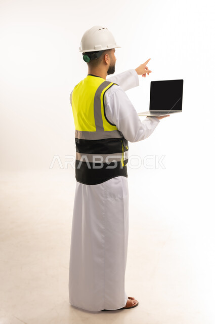 متابعة الأعمال عن بعد، الإشارة باصبع السبابة إلى مكان ما بعيد، استخدام التكنولوجيا الحديثة والمتطورة في الإشراف على العمل، بورتريه من الخلف لمهندس عربي خليجي عماني يرتدي الدشداشة والسترة وخوذة الحماية يمسك الحاسوب المحمول، الجسم بالطول الكامل، خلفية بيضاء