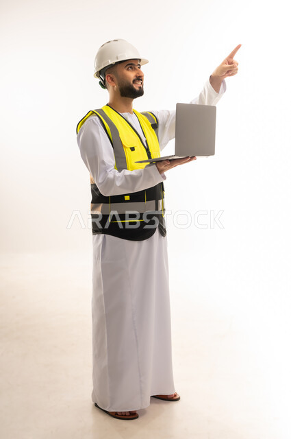 الإشارة باصبع السبابة إلى مكان ما والنظر إليه، استخدام التكنولوجيا الحديثة والمتطورة في الإشراف على العمل، بورتريه لمهندس عربي خليجي عماني يرتدي الدشداشة والسترة وخوذة الحماية يمسك الحاسوب المحمول ويقوم بمتابعة أعماله عن بعد، الجسم بالطول الكامل، خلفية بيضاء