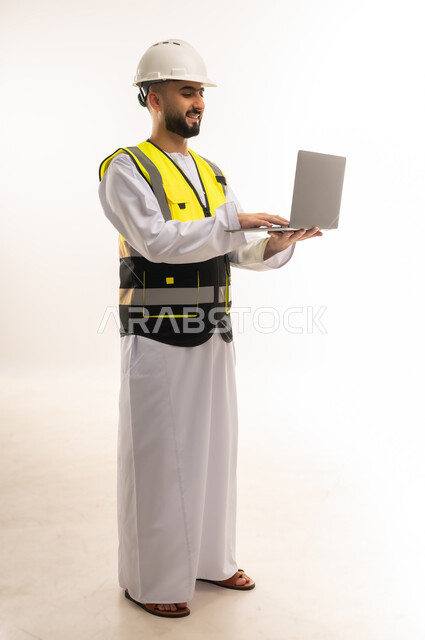 Reliance on modern technologies in the engineering sector, use of advanced technology in supervising work, portrait of an Arab Gulf Omani engineer wearing a dishdasha, jacket and protective helmet, holding a laptop and following up on his work remotely, full-length body, white background
