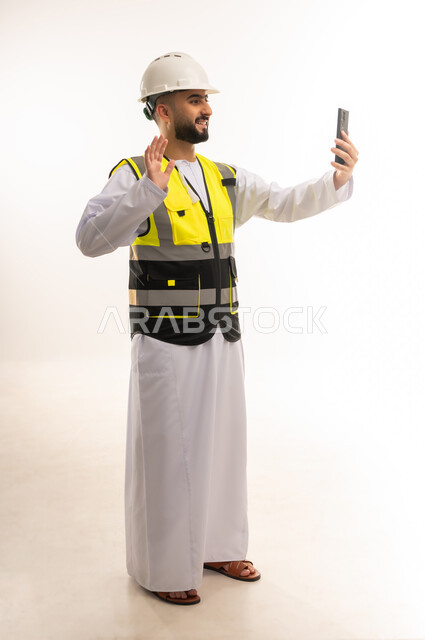Using modern technologies in the engineering sector, making video calls via mobile phone, integrating advanced technology into work, portrait of an Arab Gulf Omani engineer wearing a dishdasha, jacket and protective helmet holding a mobile device in his hand with gestures of happiness, full body length, white background