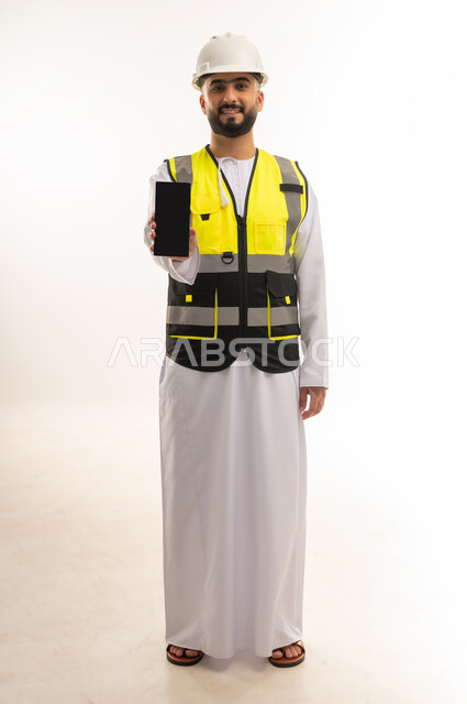 مفهوم الهندسة المعمارية، عرض شاشة سوداء فارغة عبر الهاتف المحمول، استخدام الأجهزة التقنية بالإشراف على العمل، دمج التكنولوجيا بالمجال الهندسي، بورتريه لمهندس عربي خليجي عماني يرتدي الدشداشة والسترة وخوذة الحماية يمسك الجوال، الجسم بالطول الكامل، خلفية بيضاء
