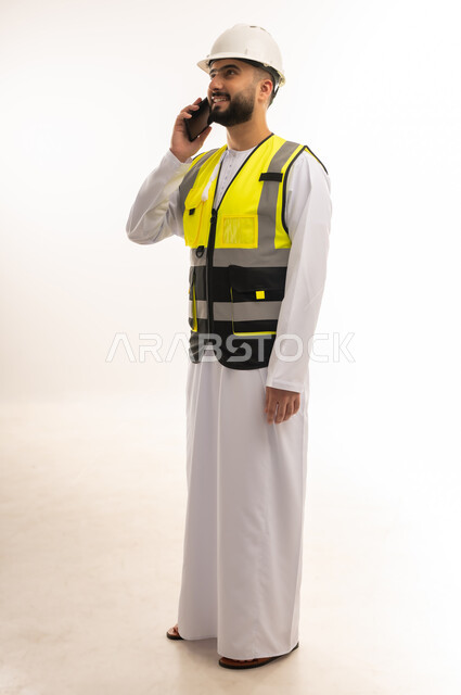 إجراء المكالمات الهاتفية باستخدام الهاتف المحمول، الاعتماد على التقنيات الحديثة بالقطاع الهندسي، دمج التكنولوجيا المتطورة بالعمل، بورتريه لمهندس عربي خليجي عماني يرتدي الدشداشة والسترة وخوذة الحماية يمسك جهاز الجوال بيده، الجسم بالطول الكامل، خلفية بيضاء