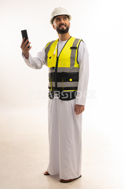 Holding a mobile phone and looking somewhere with merging gestures, using modern technologies in the engineering sector, integrating advanced technology into work, portrait of an Arab Gulf Omani engineer wearing a dishdasha, jacket and protective helmet holding a mobile phone in his hand, full body, white background