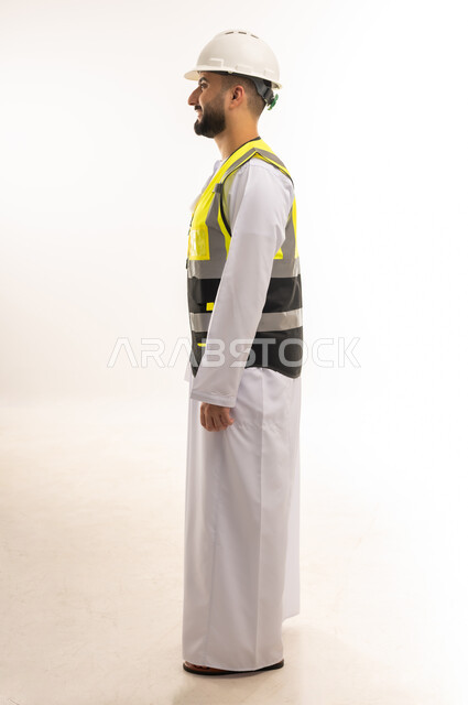 Standing straight with self-confident expression, working in architectural engineering fields, professions and jobs for young people in the Sultanate, side portrait of an Arab Gulf Omani engineer wearing a dishdasha, jacket and protective helmet looking ahead with gestures of concentration, full body length, white background