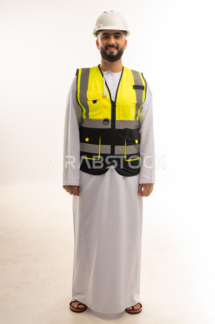 النظر إلى الكاميرا بإيماءات السعادة والبشاشة، العمل في المجالات الهندسية المعمارية، مهن ووظائف للشباب في السلطنة، بورتريه لمهندس عربي خليجي عماني يرتدي الدشداشه والسترة وخوذة الحماية يقف بشكل مستقيم بتعابير الثقة بالنفس، الجسم بالطول الكامل، خلفية بيضاء