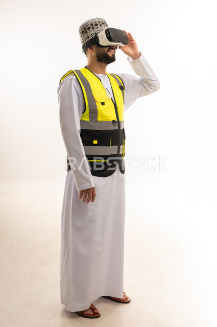 Integration and simulation of the imaginary world, fun and entertainment by watching through virtual reality glasses, love of exploration, adventure and play, portrait of an Arab Gulf Omani engineer wearing a dishdasha, a sleeve and a protective vest wearing 3D VR glasses, full body, white background