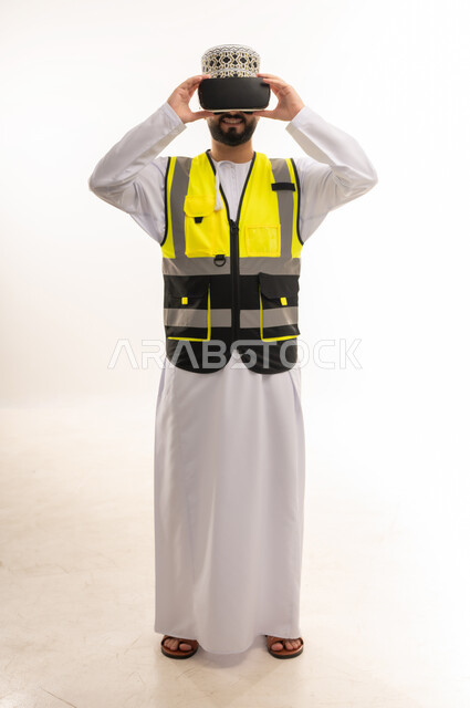 Fun and entertainment by watching through virtual reality glasses, merging and simulating the imaginary world, love of exploration, adventure and play, portrait of an Arab Gulf Omani engineer wearing a dishdasha, a sleeve and a protective vest, wearing 3D VR glasses, full body, white background
