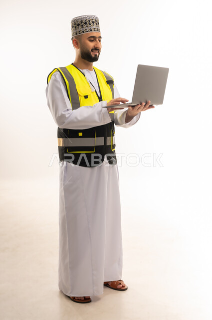 Reliance on modern technologies in the engineering sector, auditing and supervising work using advanced technology, portrait of an Arab Gulf Omani engineer wearing a dishdasha, a sleeve and a protective vest, holding a laptop and following up on his work remotely, full-length body, white background
