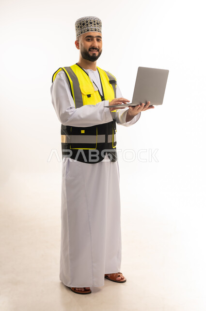 الإشراف على العمل باستخدام التكنولوجيا المتطورة، الاعتماد على التقنيات الحديثة بالقطاع الهندسي، بورتريه لمهندس عربي خليجي عماني يرتدي الدشداشة والكمه وسترة الحماية يمسك الحاسوب المحمول ويقوم بمتابعة أعماله عن بعد، الجسم بالطول الكامل، خلفية بيضاء