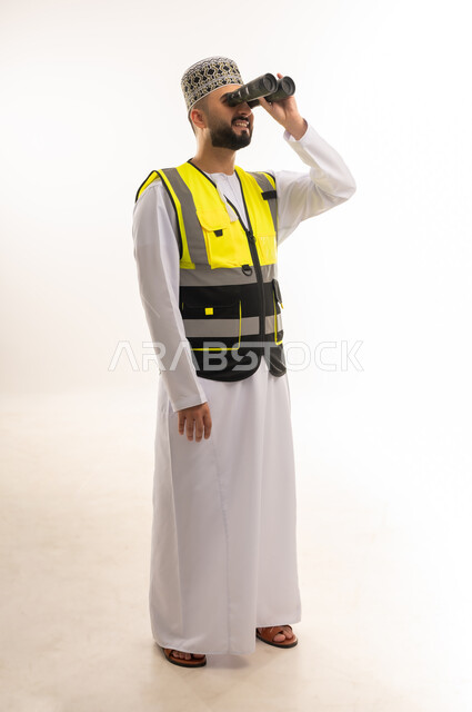 محاولة مشاهدة الاشياء البعيدة بشكل واضح، حب الاستكشاف والمغامرة، التأمل والرؤية المستقبلية، بورتريه لمهندس عربي خليجي عماني يرتدي الدشداشة والكمه وسترة الحماية يمسك بيديه منظار ثنائي العينين وينظر فيه بإيماءات البشاشة والتركيز، الجسم بالطول الكامل، خلفية بيضاء