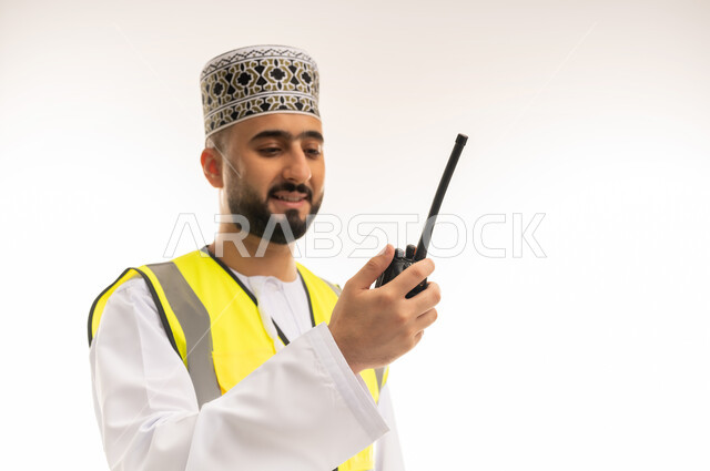 التواصل مع العمال بموقع الإنشاءات بشكل مباشر، متابعة الأعمال عن بعد باستخدام التكنولوجيا المتطورة، الاعتماد على التقنيات الحديثة بالقطاع الهندسي، بورتريه مقرب لمهندس عربي خليجي عماني يرتدي الدشداشة والكمه وسترة الحماية يمسك الجهاز اللاسلكي بيده، خلفية بيضاء