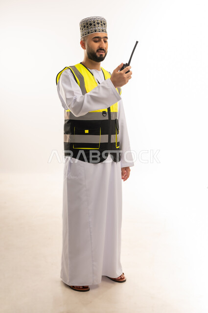 Follow-up of work remotely using advanced technology, reliance on modern technologies in the engineering sector, portrait of an Arab Gulf Omani engineer wearing a dishdasha, a sleeve and a protective vest, holding a wireless device in his hand, communicating directly with workers, full-length body, white background