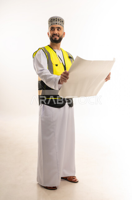 الإشراف على مخططات المشاريع الهندسية، وظائف ومهن للشباب، مفهوم الانشاءات المعمارية، العمل في القطاع الهندسي، بورتريه لمهندس عربي خليجي عماني يرتدي الدشداشة والكمه وسترة الحماية يمسك المخطط بيديه وينظر لمكان ما بإيماءات التركيز، الجسم بالطول الكامل، خلفية بيضاء