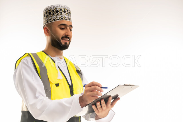 Writing the latest updates on the engineering project, taking notes, ensuring that the work is proceeding as required and correctly, close-up portrait of an Arab Gulf Omani engineer wearing a dishdasha, a sleeve and a protective jacket, holding a file in his hand and writing in it with gestures of concentration, white background