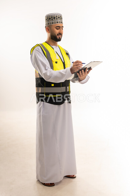 تدوين و تسجيل الملاحظات, كتابة آخر المستجدات حول المشروع، التأكد من سير الأعمال بالشكل المطلوب والصحيح، بورتريه لمهندس عربي خليجي عماني يرتدي الدشداشة والكمه وسترة الحماية يمسك الملف بيده ويكتب فيه بإيماءات التركيز، الجسم بالطول الكامل، خلفية بيضاء